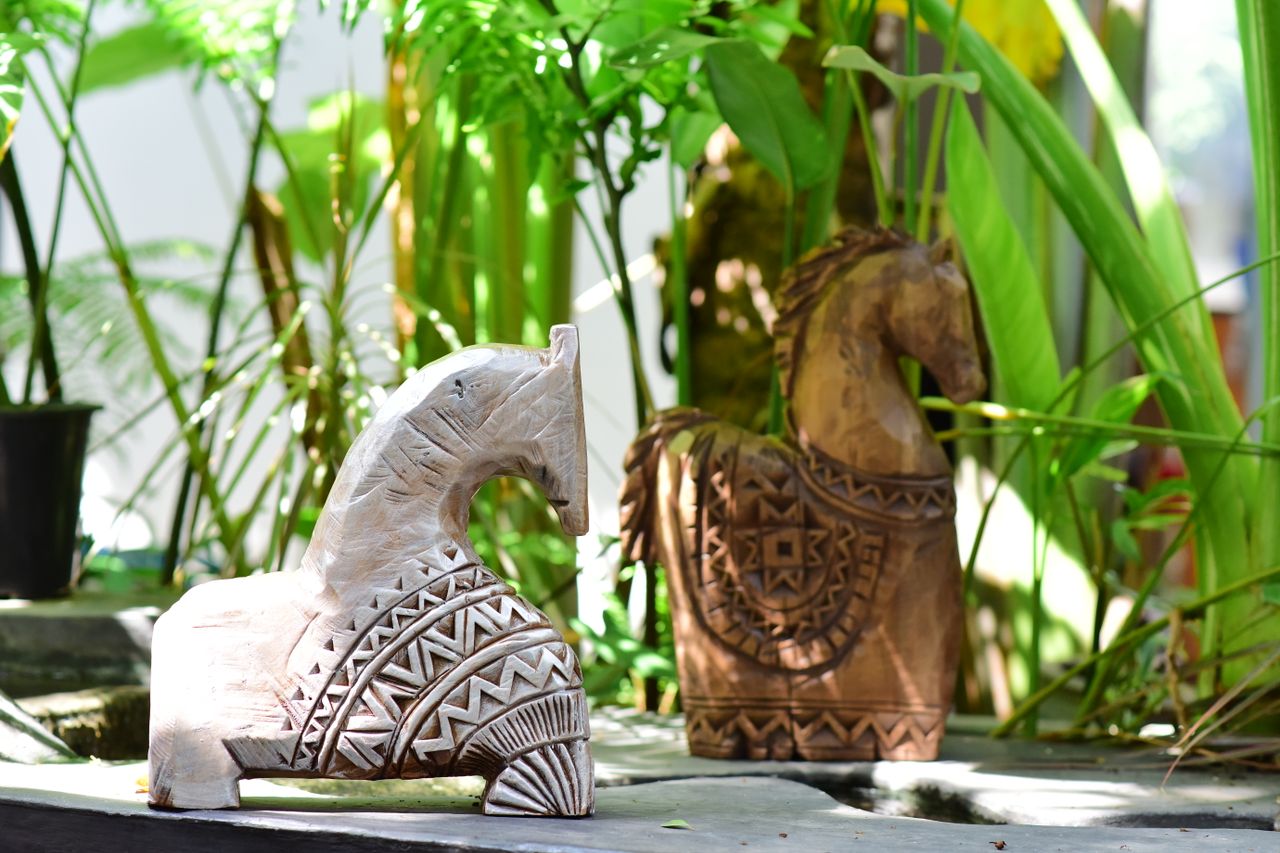 Intricately carved wooden horse sculptures displayed among vibrant green plants.