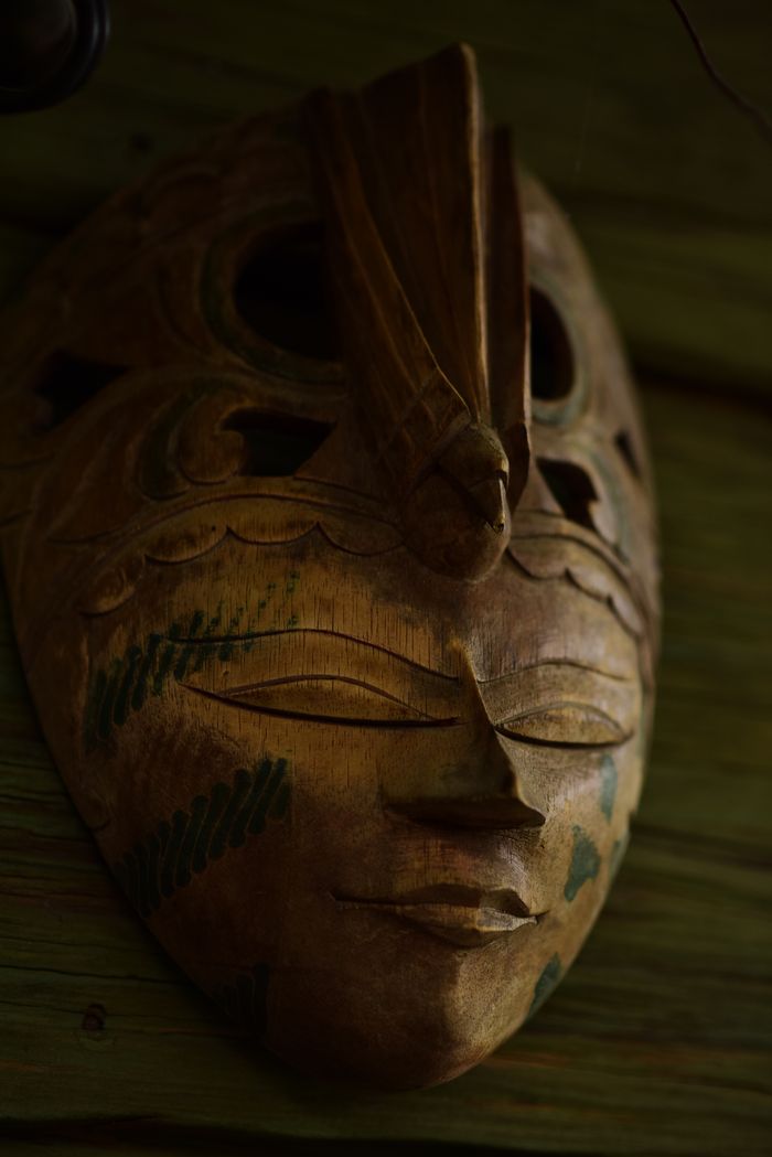 our-story A detailed tribal wooden mask with green accents displayed on a rustic wooden wall.